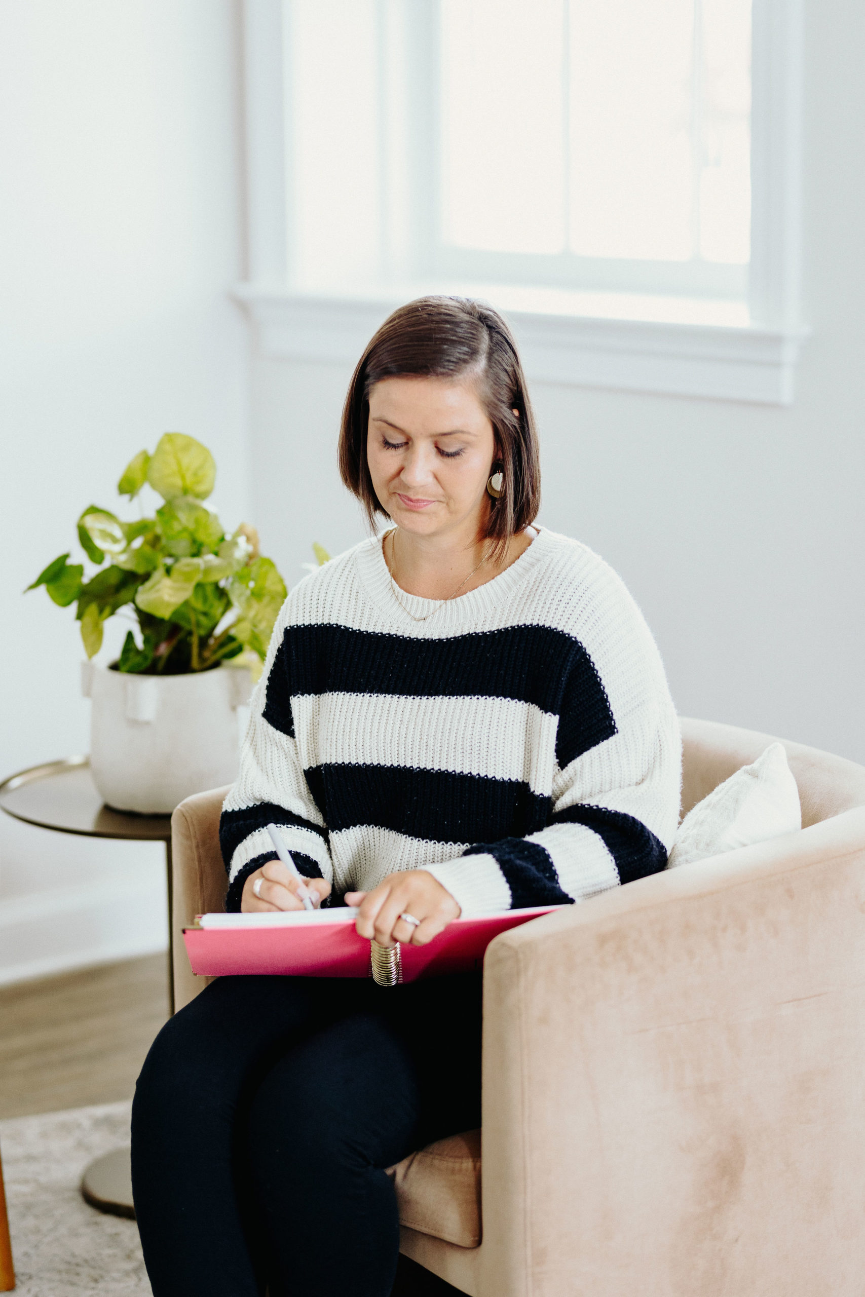 Kira from Kira Writes sitting in a cream lounge chair writing notes in her pink journal wearing a black and white striped top and black pants.